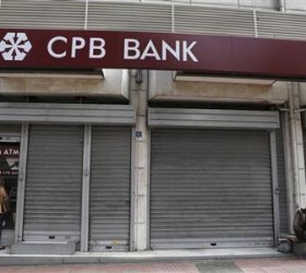 A woman makes a transaction at an ATM machine as a man begs outside a CPB branch in Athens